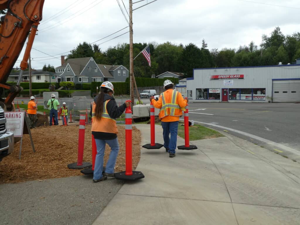 Workers carry orange traffic barriers on the Harborview Drive sidewalk where utilities are being moved for a new roundabout.