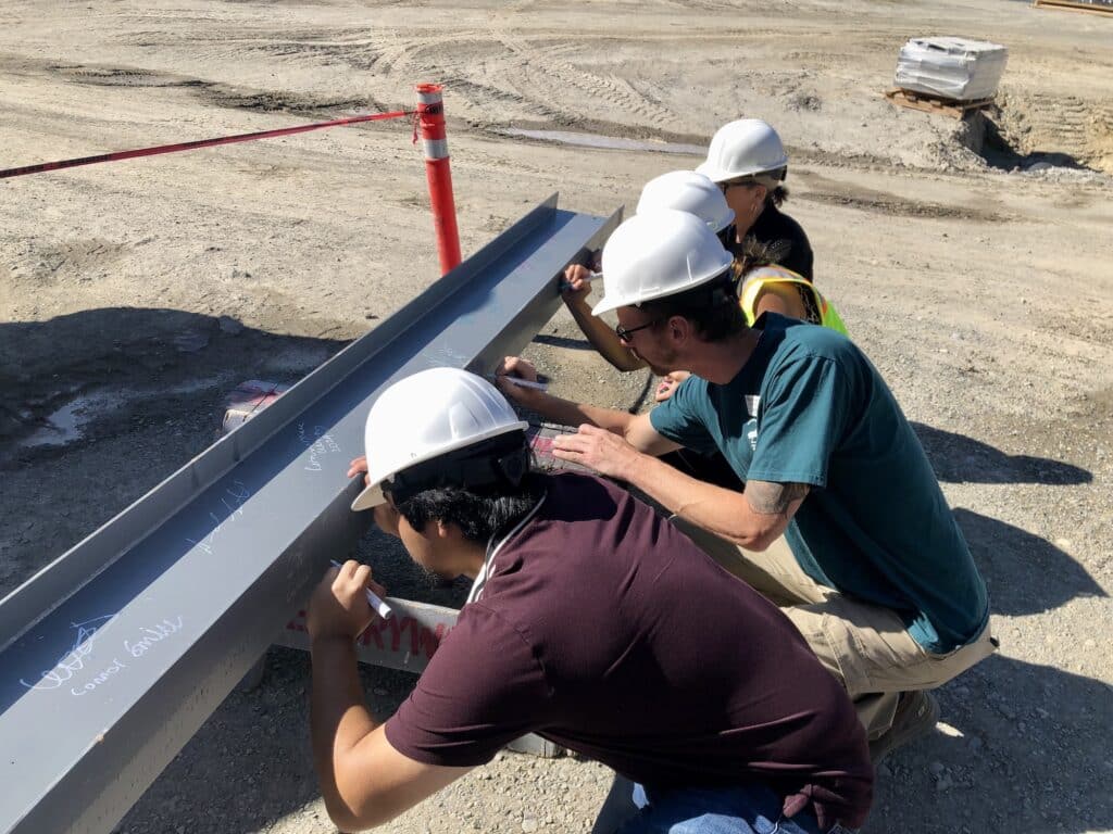 Stakeholders sign their names to the beam, including board member Billy Sehmel (green shirt) and board president Missy Hill (far end).